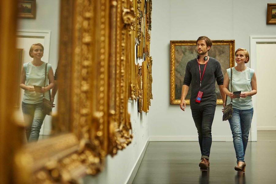 Ein Mann und eine Frau laufen in einem Museum an einer Wand vorbei. An der Wand hängen unterschiedliche Bilder mit einem Rahmen aus Gold.