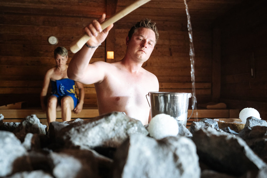 Ein Mann steht in einer Sauna und gießt Wasser über die heißen Steine. Im Hintergrund sitzt eine Frau.