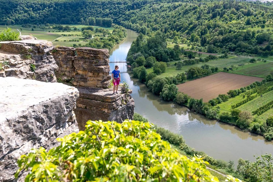 Auf einem großen Felsen überhalb des Neckars steht eine Person und genießt die Aussicht über den Fluss, die Wiesen und Weinberge.