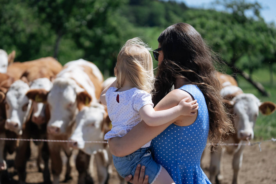 Eine Frau hält ein Kind auf dem Arm. Gemeinsam schauen sie sich die Kühe an die auf der Weide stehen.